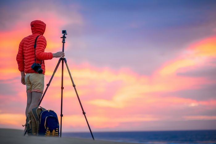 man in a red jacket holding a tripod with a camera for doing landscape photography